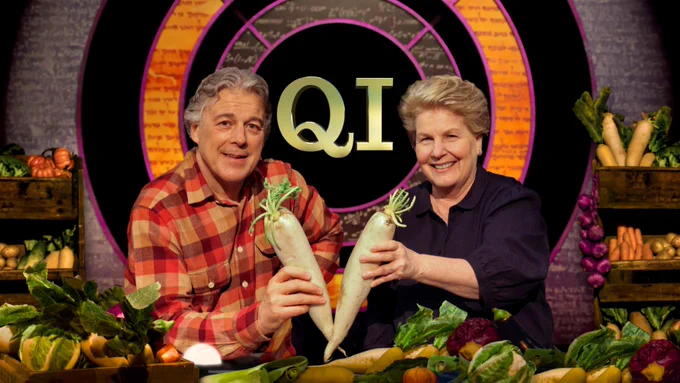 Sandi and Alan on the QI set surrounded by vegetables. They are holding two vegetables together so that they make a V shape.