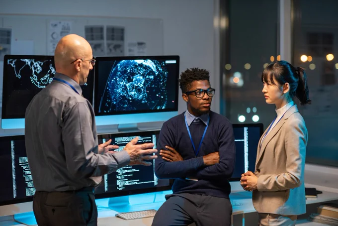 Three persons are discussing something in front of a number of computer screens. From left to right there is a man in grey shirt and black trousers and glasses, a younger man in blue swether, light blue shirt and tie. He is also wearing glasses. Lastly its a young woman in beige suit and white polo. 