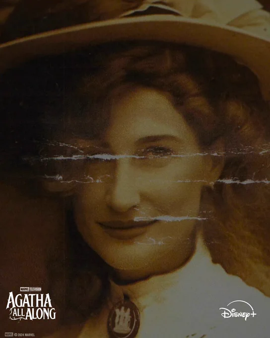 A sepia toned image of Agatha Harkness dressed in fashion from the 1900s. Her hair is curled around her face, with a large hat on top. She is wearing a white shirt with a tall collar and a brooch in the middle. The image has creases through the middle. 