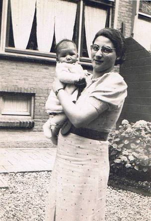 Vintage black-and-white photo of a woman holding a baby in front of a house with curtained windows and plants.