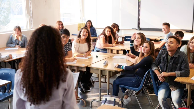 Rear view of female high school teacher standing at front of class teaching lesson.