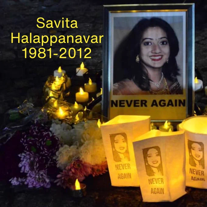 Savita Halappanavar 1981-2012  #NeverAgain  image shows candles, lanterns with Savita's face and  bunches of flowers in front of a portrait of Savita. 