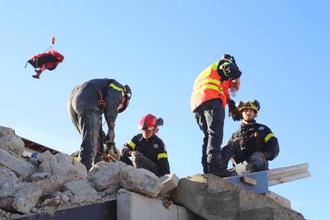 4 sapeurs-sauveteurs pendant l'exercice ; en fond, une victime se fait hélitreuiller avec un sauveteur