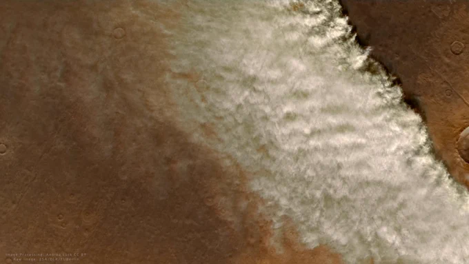 This image shows a Martian dust storm captured from orbit by ESA's Mars Express. It reveals a sweeping cloud of dust moving across the reddish-brown Martian surface. The dust storm is large, stretching over a significant portion of the landscape, and is shown as a dense, white cloud-like formation. The terrain below the storm has visible craters, characteristic of Mars’ rocky, desert-like surface.