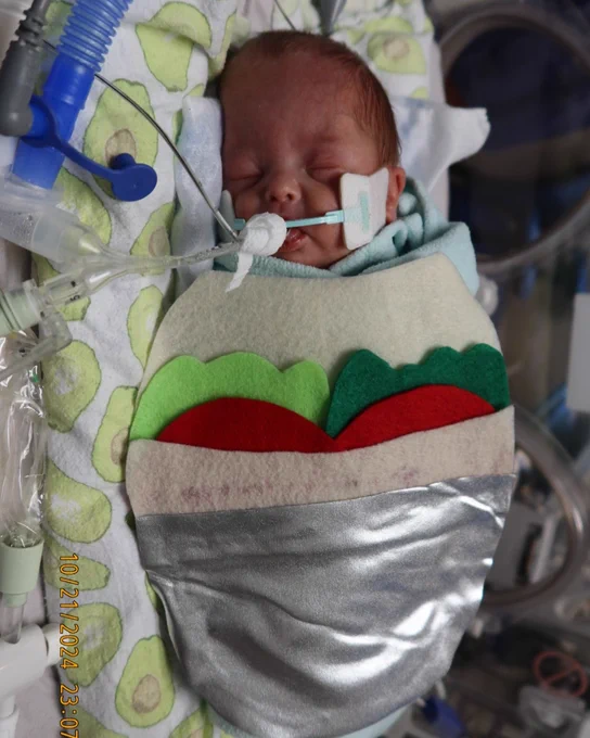 Newborn baby wrapped in a colorful blanket designed to look like a burrito, resting in a hospital incubator with a breathing tube.
