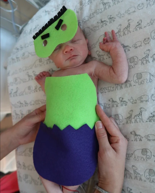 A newborn baby dressed in a homemade costume resembling Frankenstein, being held gently against a backdrop of fabric featuring various animal illustrations.