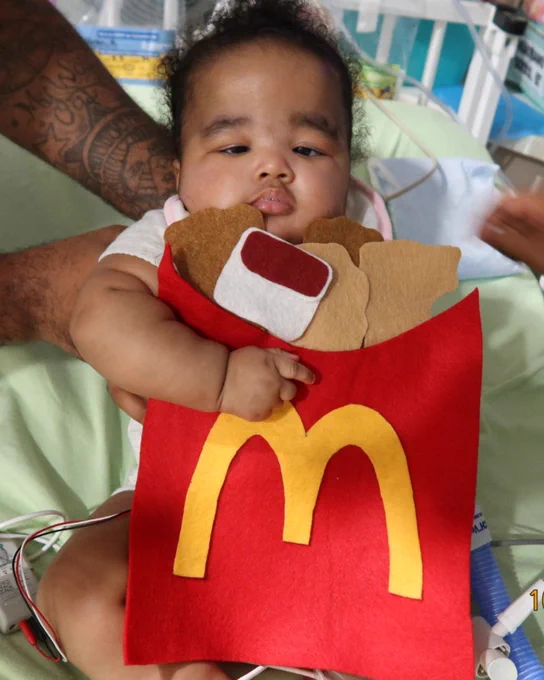 Child in hospital bed holding a McDonald's themed stuffed toy, with adult's arm visible to the side.