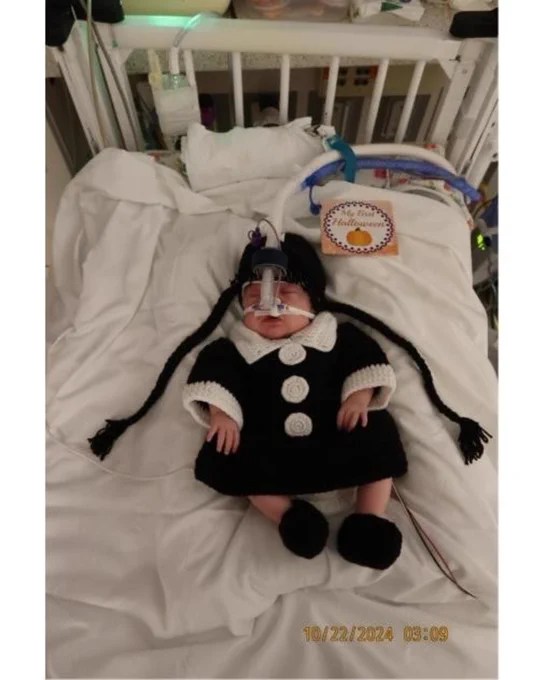 Infant in a hospital crib dressed in a black and white outfit with a pumpkin decoration nearby.