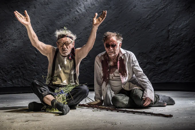A man wearing glasses with eyes attached sits on the floor next to a man with no eyes. 