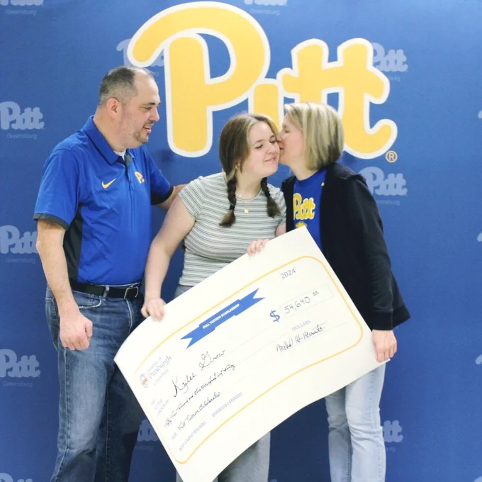 Student posing with family members, one of whom is kissing the student's cheek. Student is holding large novelty check for Full-Tuition Scholarship. Pitt-Greensburg script backdrop in the background.