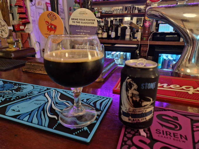 A black can stands next to a large goblet glass half full of back liquid with a light foam head. They are both on colourful bar runners atop a wooden bar with handpull behind 
