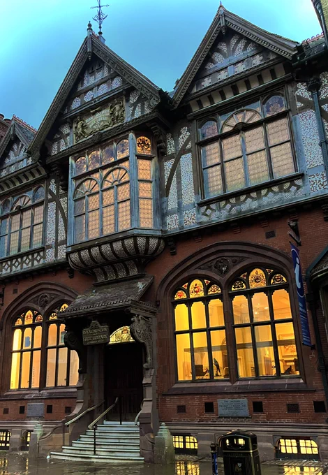 The front of The Beaney, a Tudor style building with large arched windows a central staircase and decorative woodwork. An orange light glows from the large windows and is reflected on the  wet pavement.