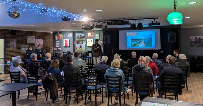 A group of people sitting on chairs in a room watching a presenter displaying a PowerPoint presentation about the Pevensey Bay to Eastbourne Scheme. 