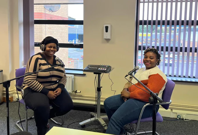 Mena from Merthyr Valleys Homes and Evelyn from Tai Pawb are sat in an office with windows behind him, they are recording a podcast and there are microphones and a sound deck in-between them. They are both wearing jumpers in the chilly weather.