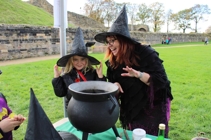 A young visitor wearing a witch's hat and smiling, stood next to our wonderful witch, behind a cauldron at Pontefract Castle
