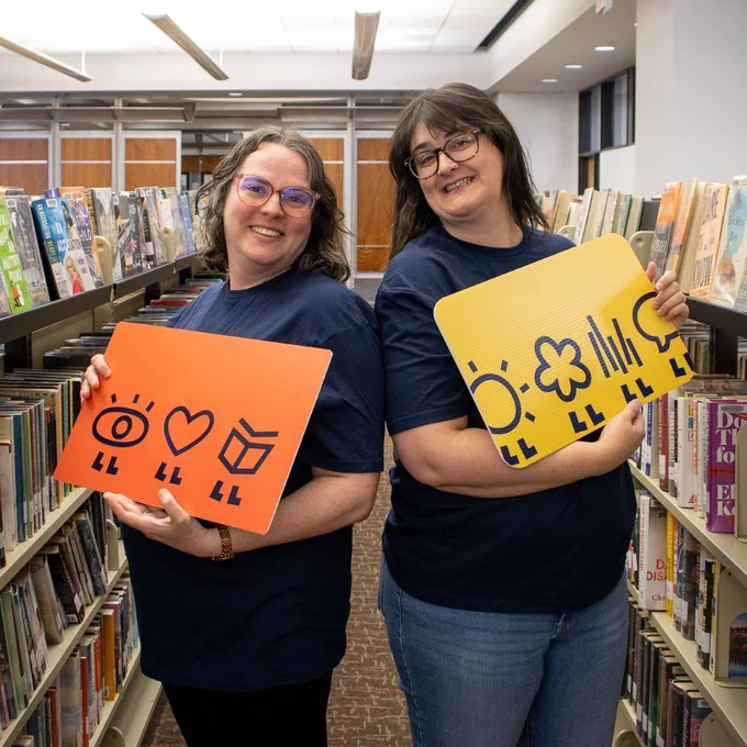 2 Librarians back to back between bookshelves holding large replicas of London Public Library, Library cards.