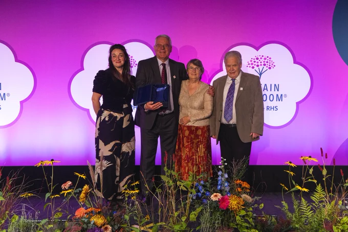 Four people on a stage with the Britain in Bloom logo behind them and floral arrangements in front. They are all smiling and one of the men is holding an award