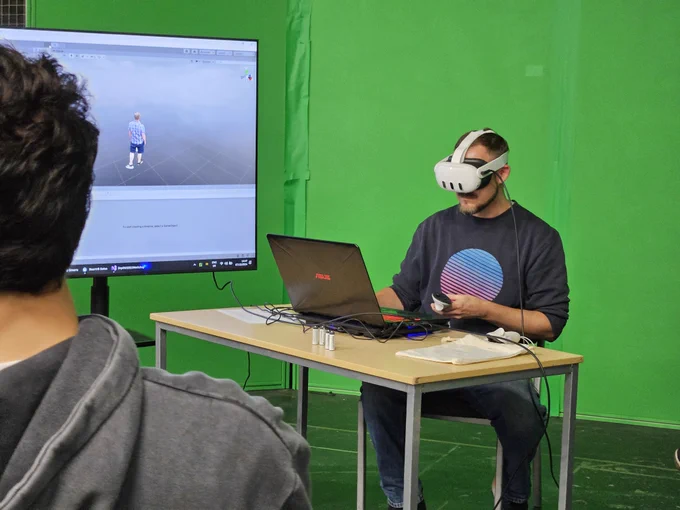 Man wearing a VR headset sits at a table with his laptop in front of a green screen