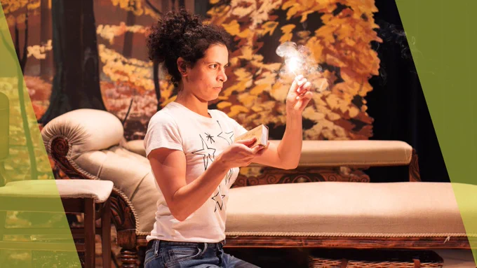 Production photo from AUTUMN - Rebecca Banatvala, playing Elisabeth, lights a match. Behind her is a chaise longue - and a painting of an Autumnal scene.