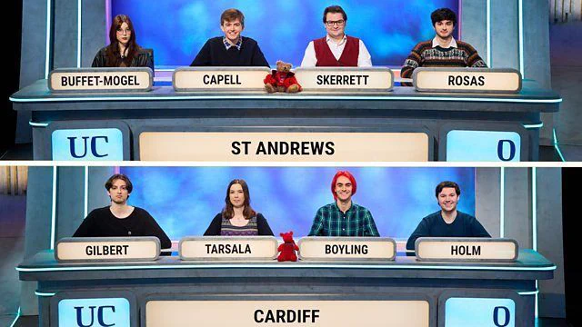 University Challenge. St Andrews University team: Buffet-Mogel; Capell; Skerrett and Rosas. They have a small bear mascot on their desk. Cardiff University team: Gilbert; Tarsala; Boyling, and Holm. They have a small dragon mascot on their desk.