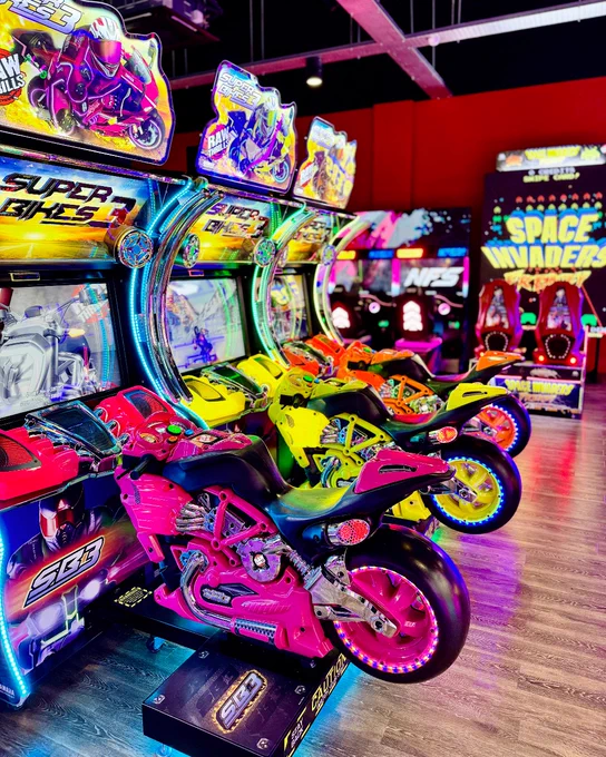 Row of colourful Super Bikes arcade racing games inside a vibrant arcade room, featuring other illuminated game machines in the background.