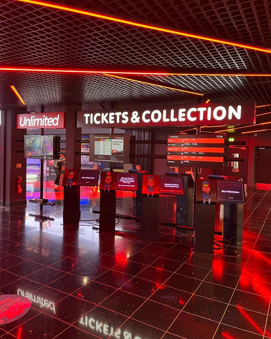 A brightly lit interior of a Cineworld cineme with "Tickets & Collection" counters, featuring multiple self-service kiosks. The setting is modern with red and black decor, prominent signage for the "Unlimited" program, and a reflective floor.