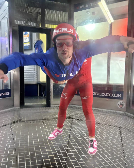 Person in a blue and red jumpsuit with iFLY logo and safety gear is indoor skydiving in a vertical wind tunnel.