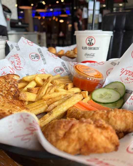 A meal from Slim Chickens, featuring crispy chicken tenders, French fries, cucumber slices, and a side of sauce, served on a branded paper with a Coca-Cola cup in the background.