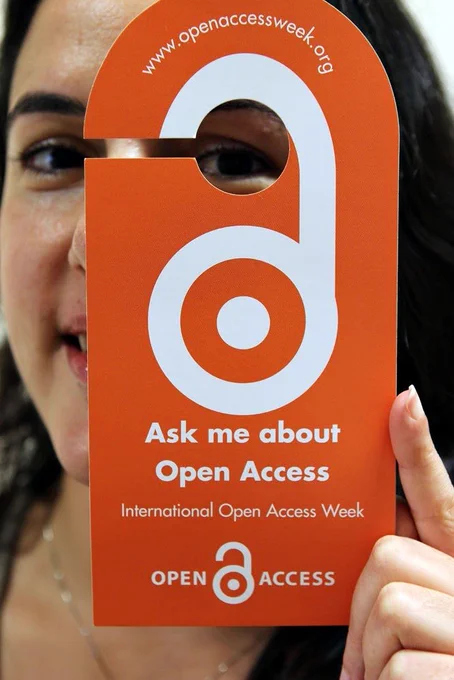 Promotional image from the International Open Access Week committee. A person holds an orange door hanger with a circular cut-out, partially covering their face. The sign reads, "Ask me about Open Access. International Open Access Week," with the Open Access logo at the bottom