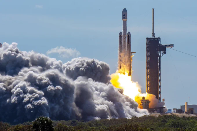 A rocket with two side boosters rides a column of flame as it lifts off the launch pad on a sunny day. Billowing clouds of steam flow from the pad.