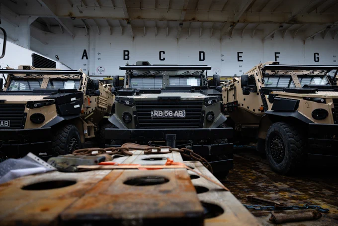 A green military vehicle sits between two beige military vehicles on a ferry.