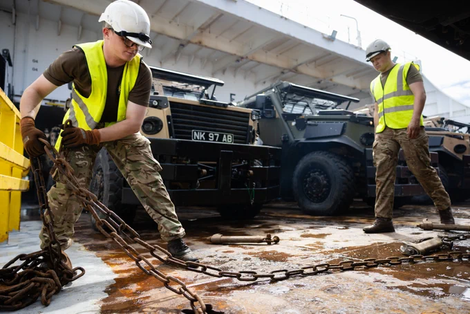 A soldier wearing a hi-vis jacket pulls a chain whilst another soldier stands on the right. There are vehicles behind them.