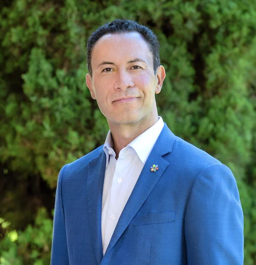 A man in a white shirt and blue suit smiling at the camera. In the background is a green bush.