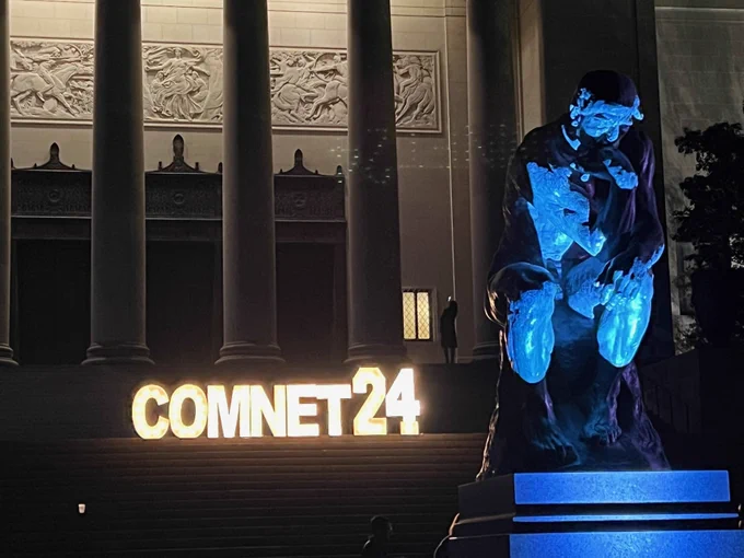 A statute of the Thinker is seen in the foreground and a lighted ComNet24 sign is seen on the steps of a museum with tall columns. 