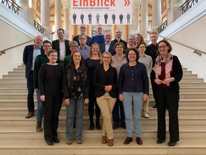 Teilnehmer:innen des Treffens stehen auf einer Treppe im Treppenhaus der BSB in München. Hinter ihnen hängt ein Banner der Fotoausstellung "Einblick. Fotografien von Volker Hinz"