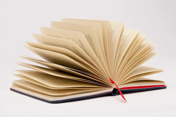 An open book with cream-colored pages is slightly fanned out against a white background. A red ribbon bookmark is visible, adding a pop of color to the otherwise neutral-toned image. The image suggests open knowledge and creativity. 