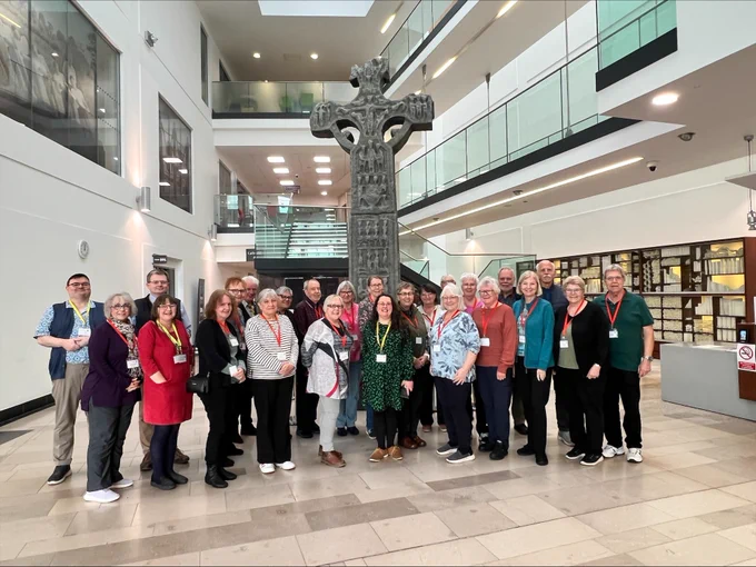 Photograph of a group of people from the Ulster Historical Foundation and their delegates standing in the PRONI atrium in front of a huge celtic cross.