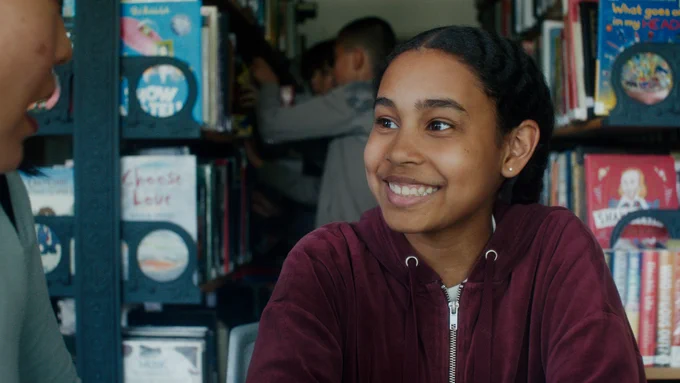 A child in a burgundy hoodie smiling and looking at an adult who is talking to them. They are in front of library stacks.