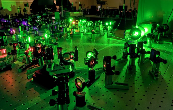 A metal table covered in holes is lit bright green by the laser beam passing through various optics and mirrors screwed into the table. In the background, a scientist is working on the setup