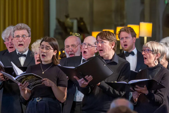 A mixed-age choir passionately sings during a performance, with members holding open music scores. The group includes men and women, some in formal black attire, while others wear more casual black outfits. The singers appear deeply engaged, their expressions reflecting the intensity of the music. The background shows soft, warm lighting from lamps, creating an intimate and focused atmosphere within a church or concert hall setting.