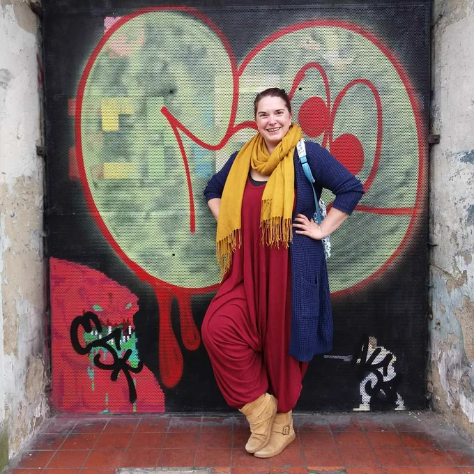 Sarah is a young white woman with tied back brown hair, standing in a doorway where the wall behind her is covered in large colourful grafitti tags, She wears long deep red baggy harem-pant jumpsuit, long blue cardigan and large mustard-yellow wraparound scarf. She stands hands on hips and smiling.