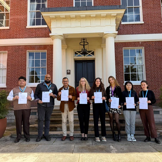 A group of eight bursary winners standing in front of the entrance to Hinxton Hall. They are each holding up a certificate. 