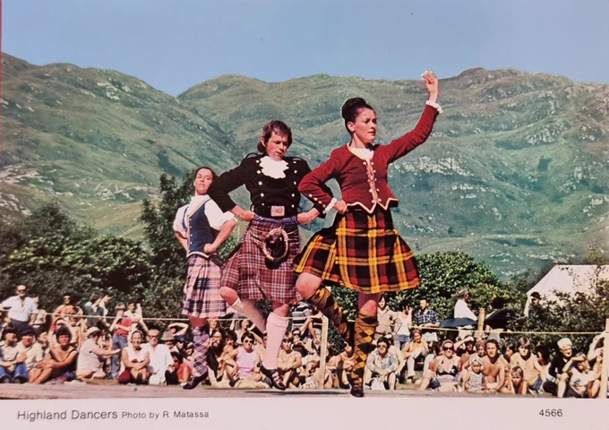 A late 20th century postcard of three dancers in kilts and waistcoats. They are mid dance on an openair dancefloor at a Highland Games or similar event. In the background is a range of hills. It appears to be a hot summer day. A large crowd looks on and some men are topless. 