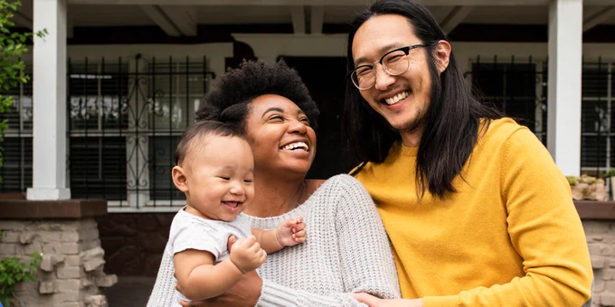A joyful family of three, including an adult holding a smiling baby and another adult, standing in front of a house with a porch. They are all smiling warmly.