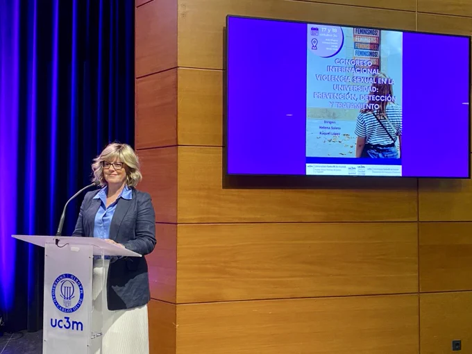 Professor Rachel Fenton stands at a white lectern with a blue logo. Behind her is a blue curtain, some orange wood panelling, and a screen showing the title of the International Congress.