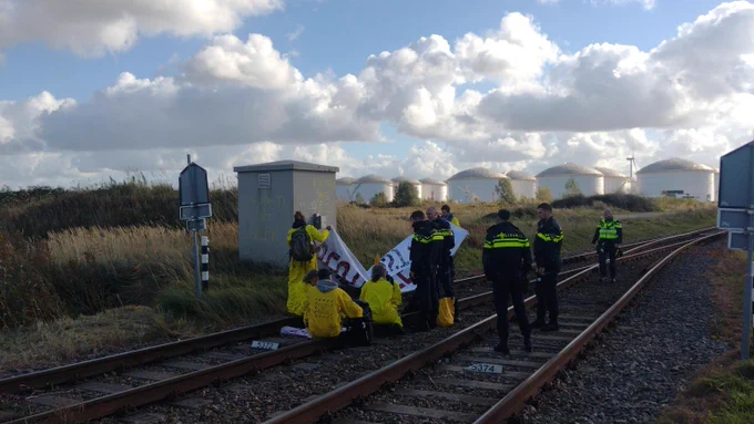 Zwei gleise, auf denen noch etwa 5 gelb bekleidete Personen sitzen. Neben ihnen stehen 5 niederländische Polizist*innen.