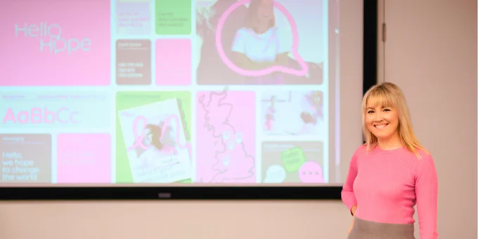 The author of the article, Ellie MacDonald, stands in front of a PowerPoint slide projection. The slide is for the community interest company that Ellie set up, HelloHope. Ellie has blonde hair just past her shoulders and she’s wearing a pink long-sleeved jumper and a grey skirt.