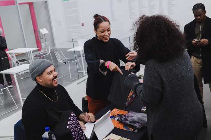 A photo of two people at a table handing out copies of Seen Journal. They are smiling, there is someone visiting the table picking up a copy. 