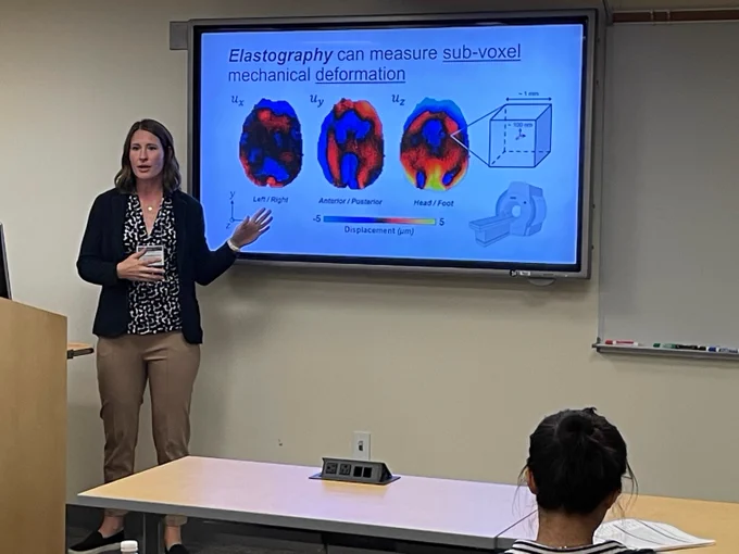 A photograph of a speaker in front of a slide with a title that reads: Elastography can measure sub-voxel mechanical deformation