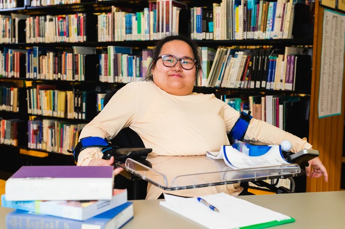 Rachel, a young woman who uses a wheelchair, poses for a photo behind a desk in a library